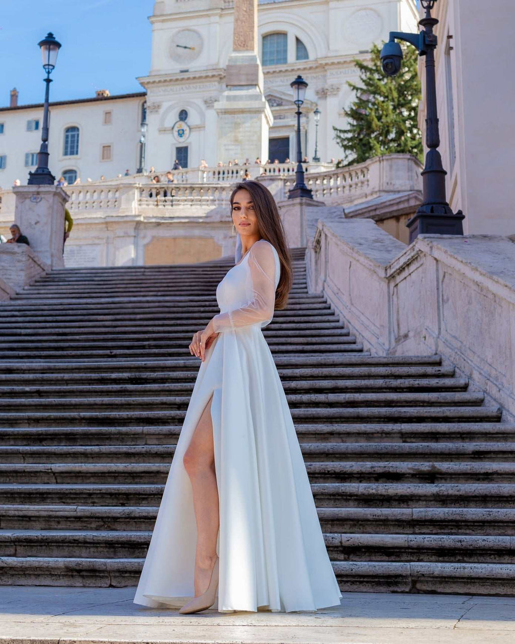 Rochie de mireasa albă lungă din satin cu șliț elegantă și decolteu frumos, fundal arhitectural.