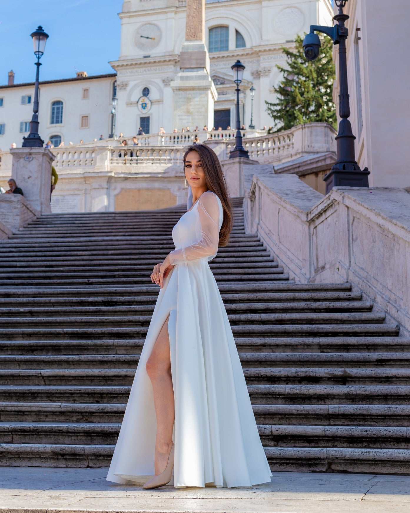 Rochie de mireasa albă lungă din satin cu șliț elegantă și decolteu frumos, fundal arhitectural.